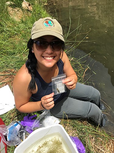 Young woman holds up bagged insect speciman while sitting next to stream