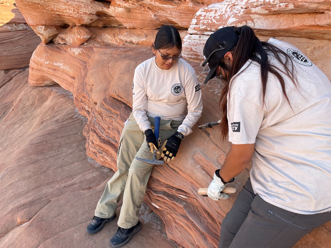 Two people remove graffiti from sandstone wall using a rock hammer and wire brush.