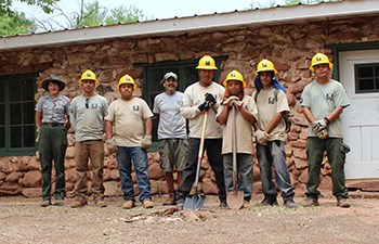 Group of young men in hardhats stand with park employees in front of historic stone building