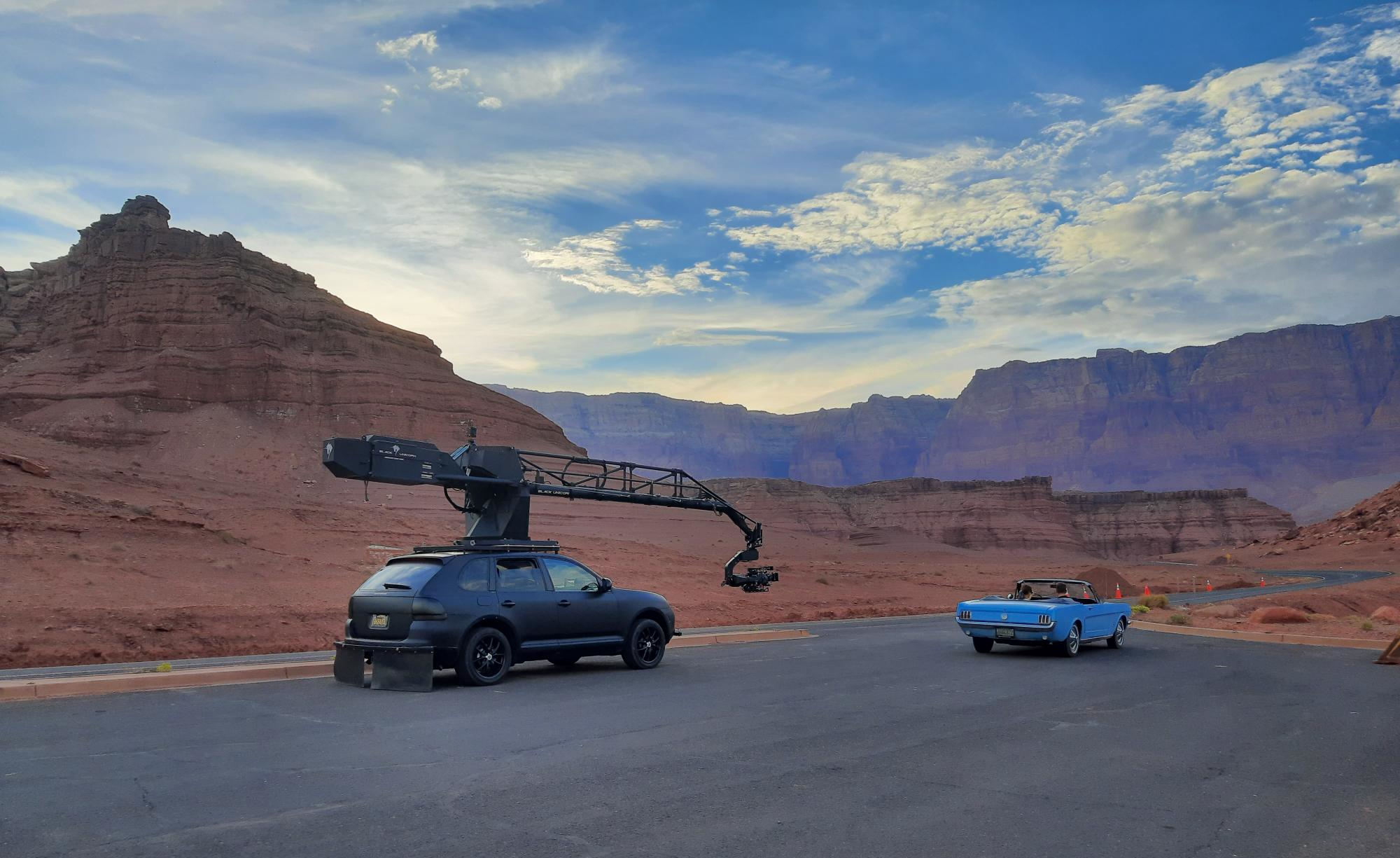 A baby blue 60s convertible drives out of a parking lot. A black car with a filming camera on a crane attached to the roof follows it.