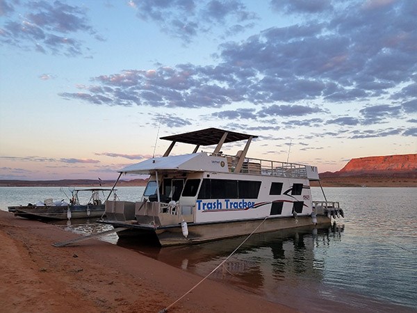 A houseboat and runabout are beached on a lake. Cliffs and clouds in the background.