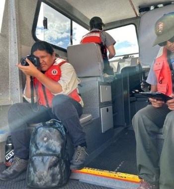 Person in lifejacket sits on a low seat in a boat, holding a camera to thier face. A similarly dressed boat driver has their back to the seat an a third ranger in a lifejacket reads a clipboard.