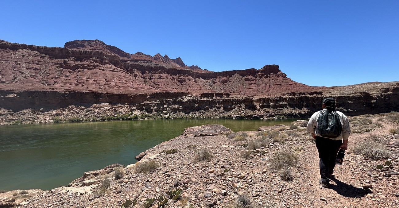 Bright future ahead! Person wearing a dark backpacka nd carrying a water bottle walks along a dusty trial next to a clear river in a sandstone canyon