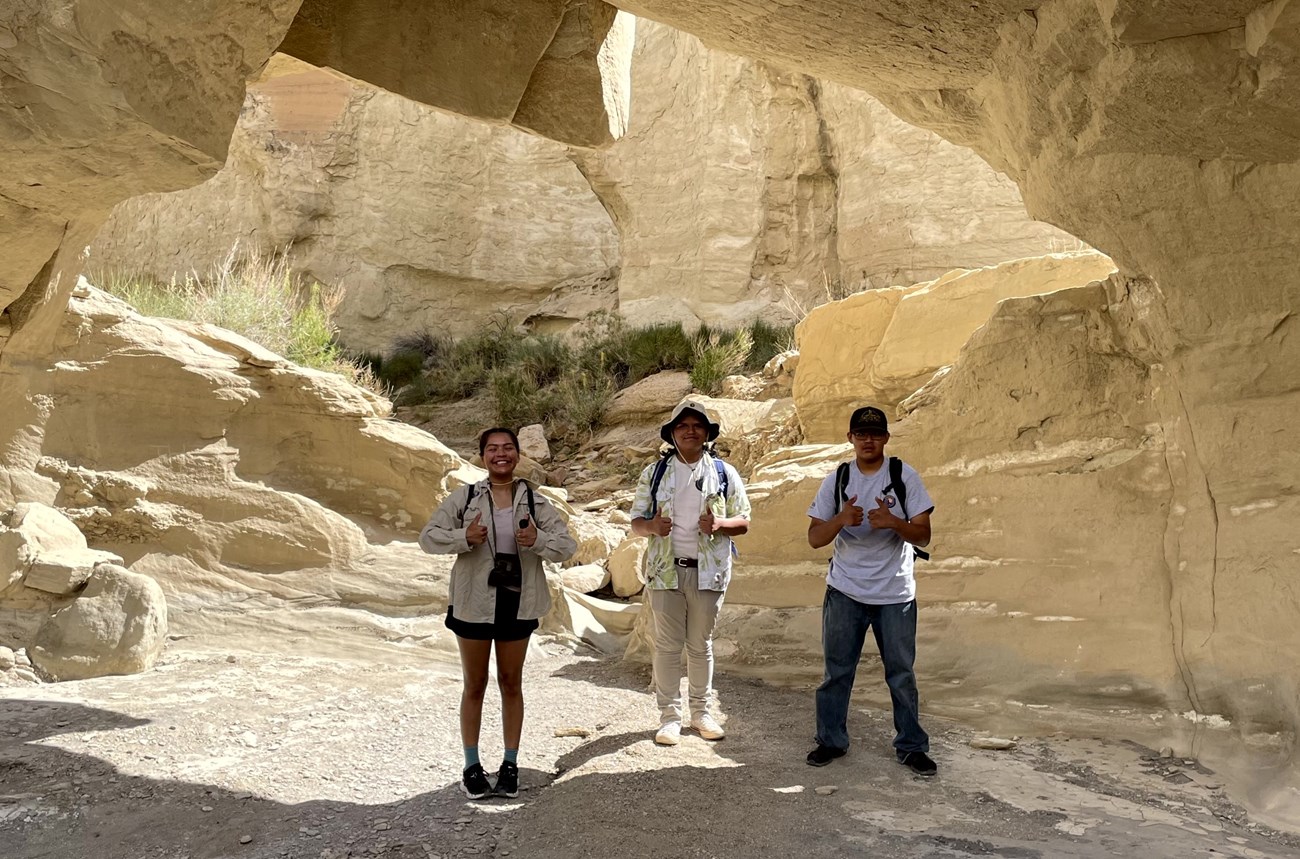 Glen Canyon 2023 Interns Three people stand underneath a natural sandstone arch. They are all giving double thumbs up.
