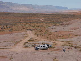 Two vehicles are parked at the end of a dirt road on a sandstone surface. A group of people stands near the trucks. Two vehicles are parked at the end of a dirt road on a sandstone surface. A group of people stands near the trucks.