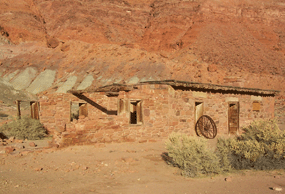 Modern image of historical bulding which blends into its surroundings. Half of the building is crumbling, with only wooden window fromes showing. A wagon wheel leans against the building.