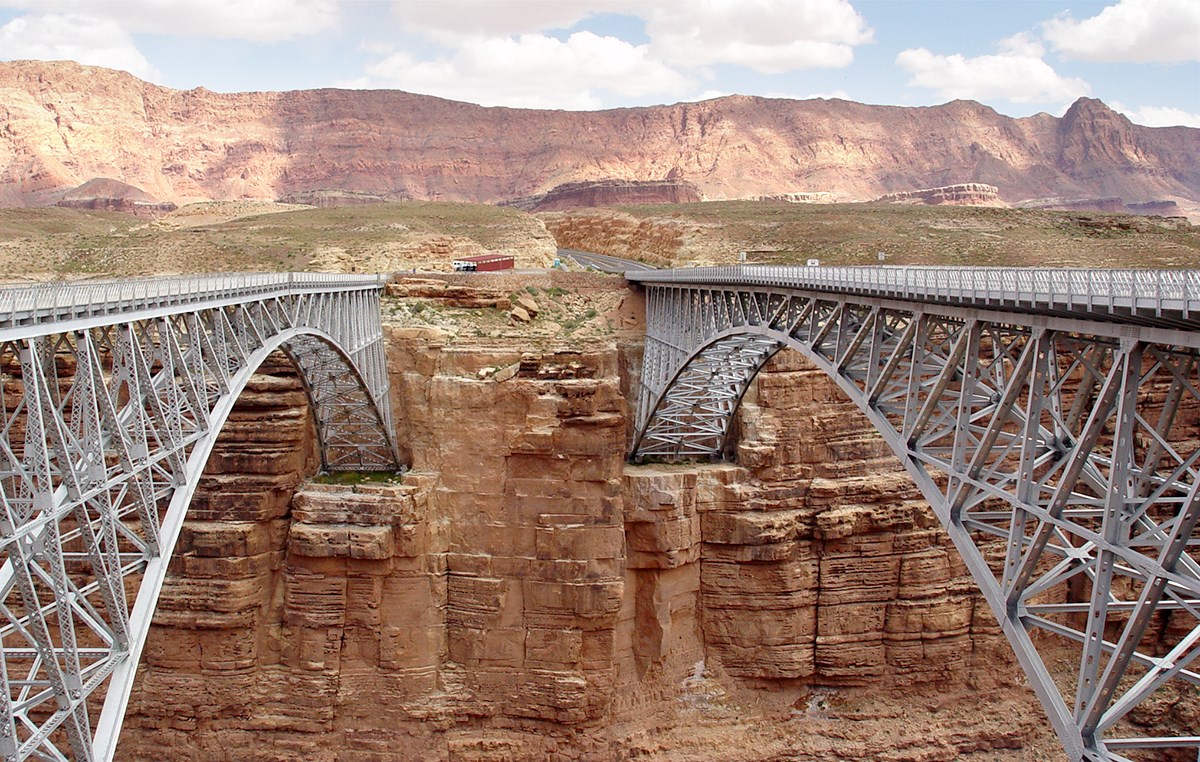 Two almost identical steel-arch bridges span the canyon. A parking area is on the other side.