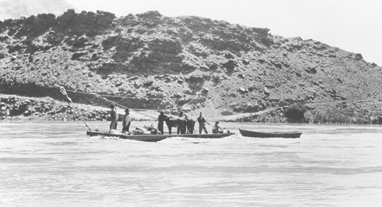 In a historic pohoto, six men, a horse, and gear ride a cable ferry across the Colorado River. They drag a small boat behind them.