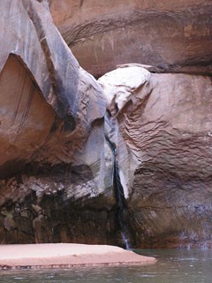 A natural feature called Cathedral in the Desert. Swoopy sanstone cliffs with a natural waterfall in the center. Sandy beach at the bottom.