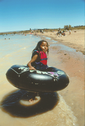 Child wearing large black inner tube and red lifejacket walks out of the shallow water onto the beach. Large swimming crowd in background.