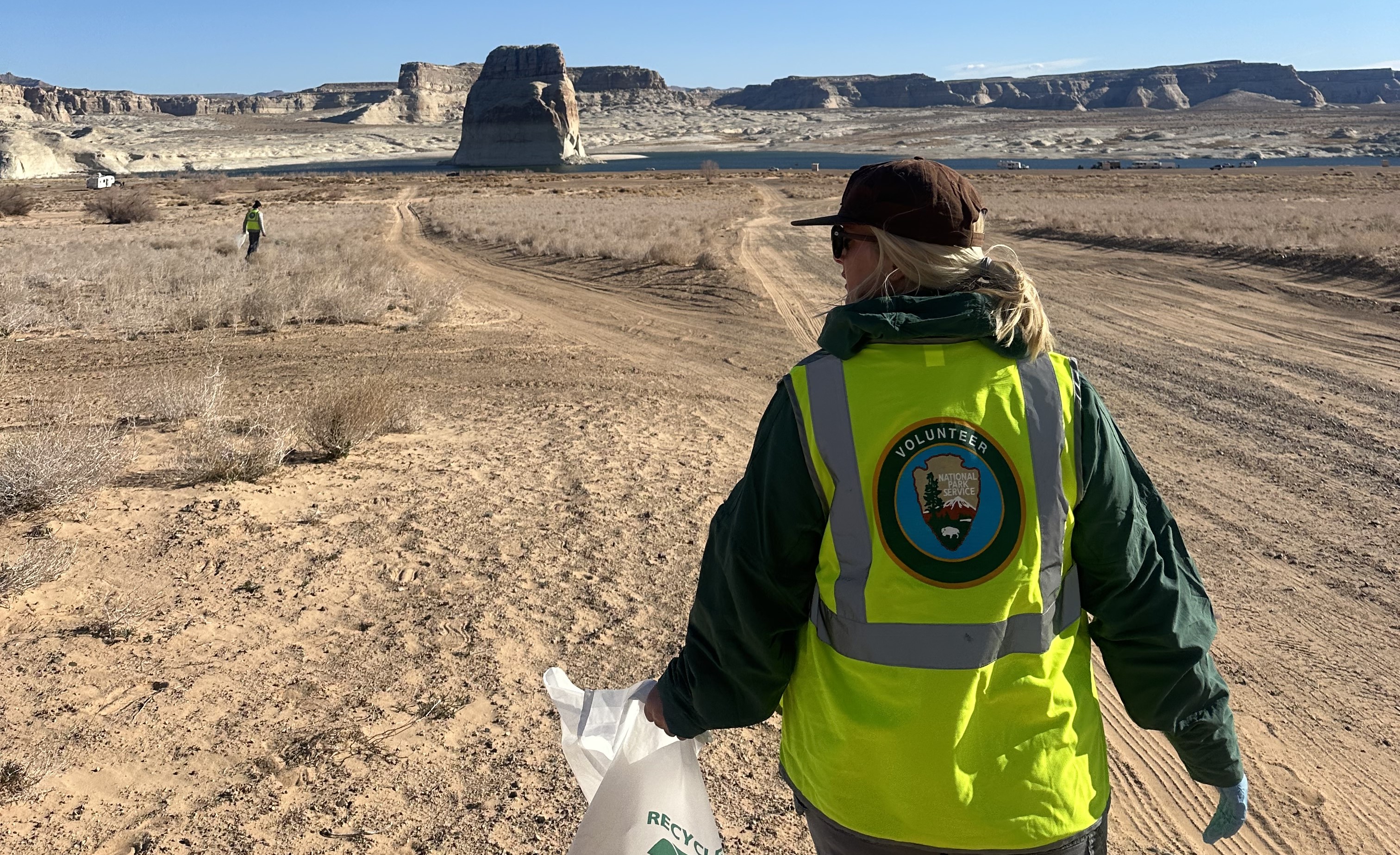 Volunteer Cleanup Person in a yellow safety vest with the NPS Volunteer logo on the back walk down a beach holding a trash bag. A large butte in a desert lake is in the background.