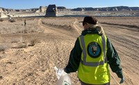 Person in a yellow safety vest with the NPS Volunteer logo on the back walk down a beach holding a trash bag. A large butte in a desert lake is in the background.