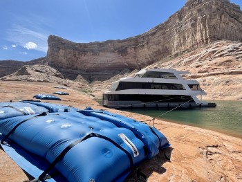 Multiple large blue bags full of water sit flat on sandstone rock ledge beach. They are tethered with ropes to a large houseboat floating on the water close by. The area is surrounded by a large canyon wall.