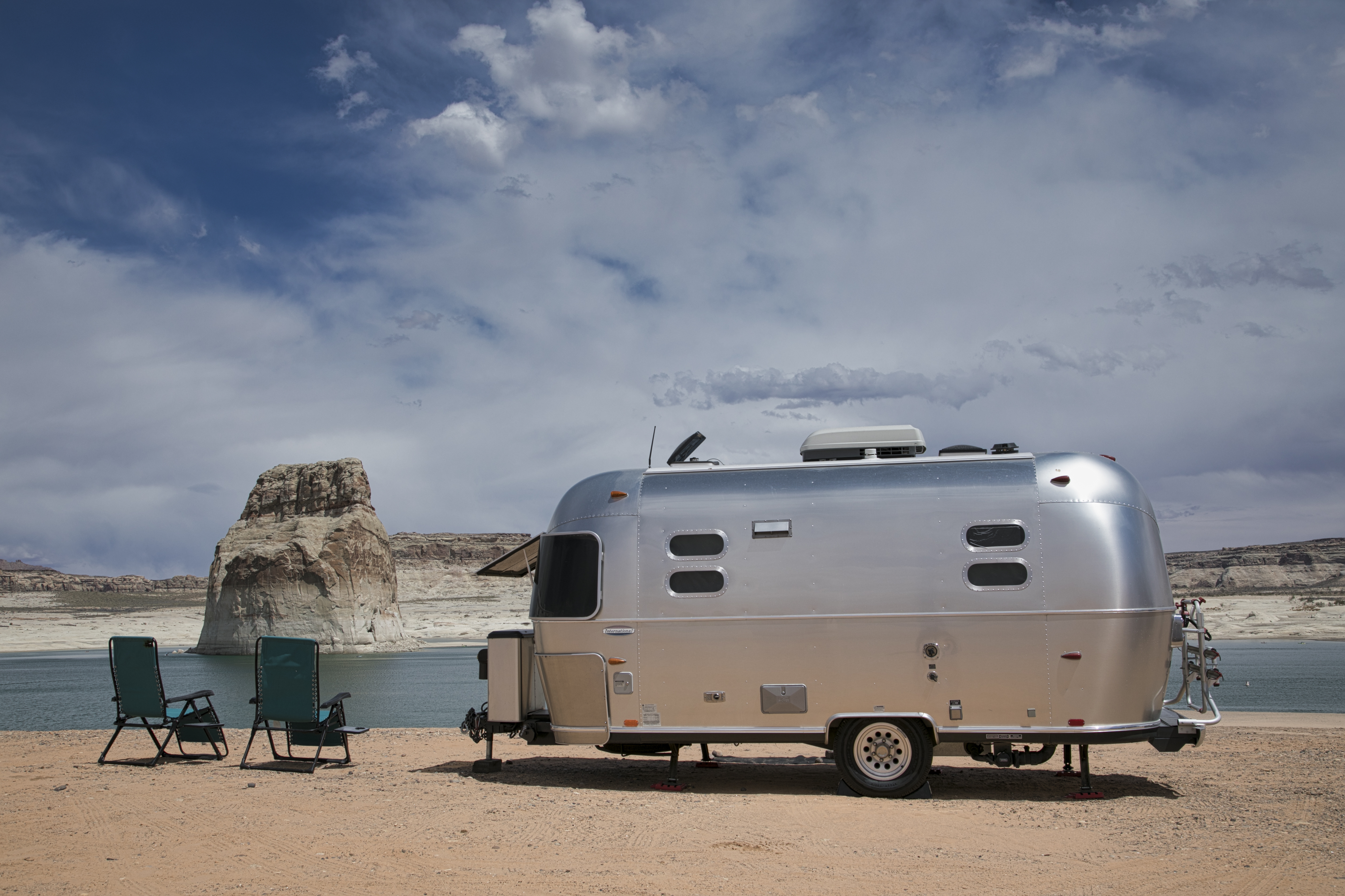 A metal camper parked on beach with two lawn chairs beside it