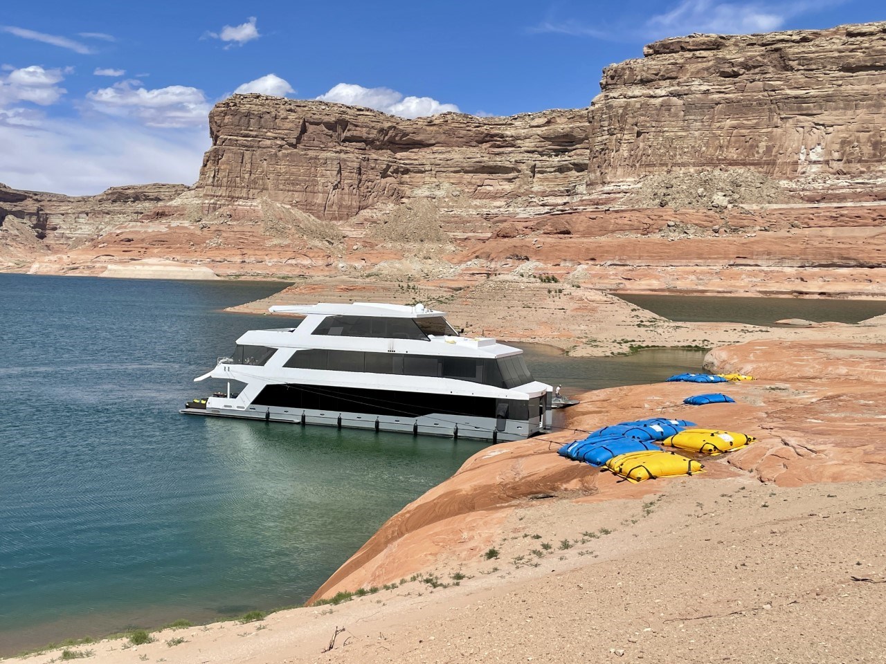 Multiple large yellow and blue bags full of water sit flat on sandstone rock ledge beach. They are tethered with ropes to a large houseboat floating on the water close by. The area is surrounded by a large canyon wall.