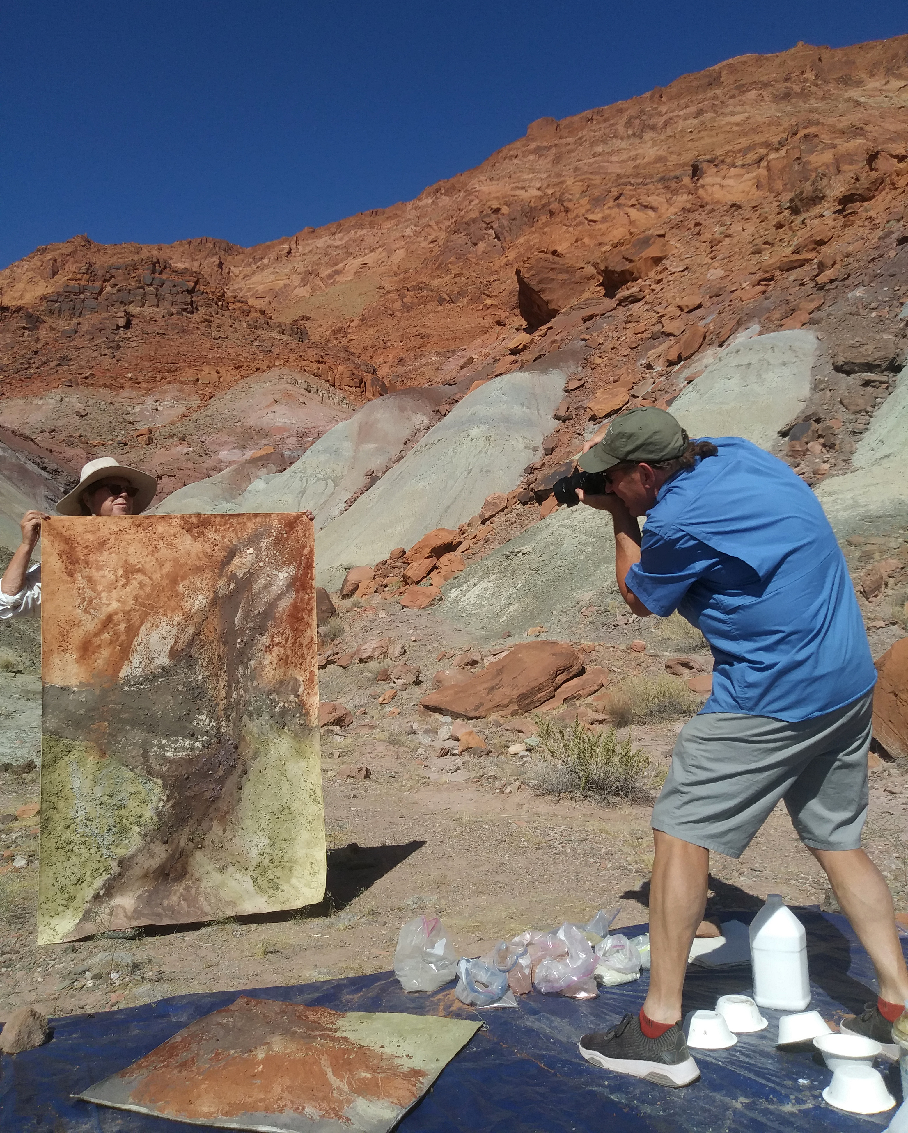 Man photographs an artist holding a canvas covered in colors and textures that match the landscape around them