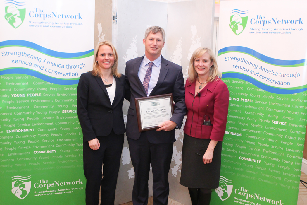 Lonnie Pilkington (center) receives Champion of the Year Award from Amy Sovocool (left) and Mary Ellen Sprenkel (right) Lonnie Pilkington holds plaque with his name on it. People in businesswear on either side of him.
