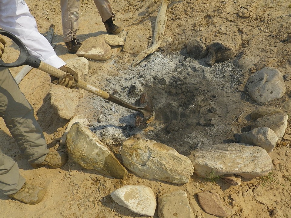 Make Sure Your Campfire is Completely Out. Ashes in fire ring on sandy area next to legs of two people. A shovel pokes at the ashes.