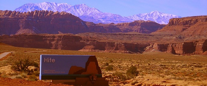 Park entrance sign at Hite surrounded by cliffs with snowcapped mountains in background
