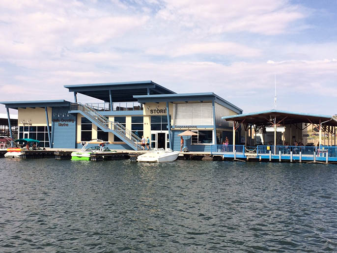 A beige structure with brown trim sits on top of a dock with boats pulled up to it. The building is labeled at Halls Crossing Store.