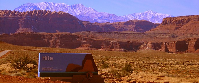 Hite District of Glen Canyon National Recreation Area Sign saying "Hite" surrounded by sandstone cliffs, snow-topped mountains