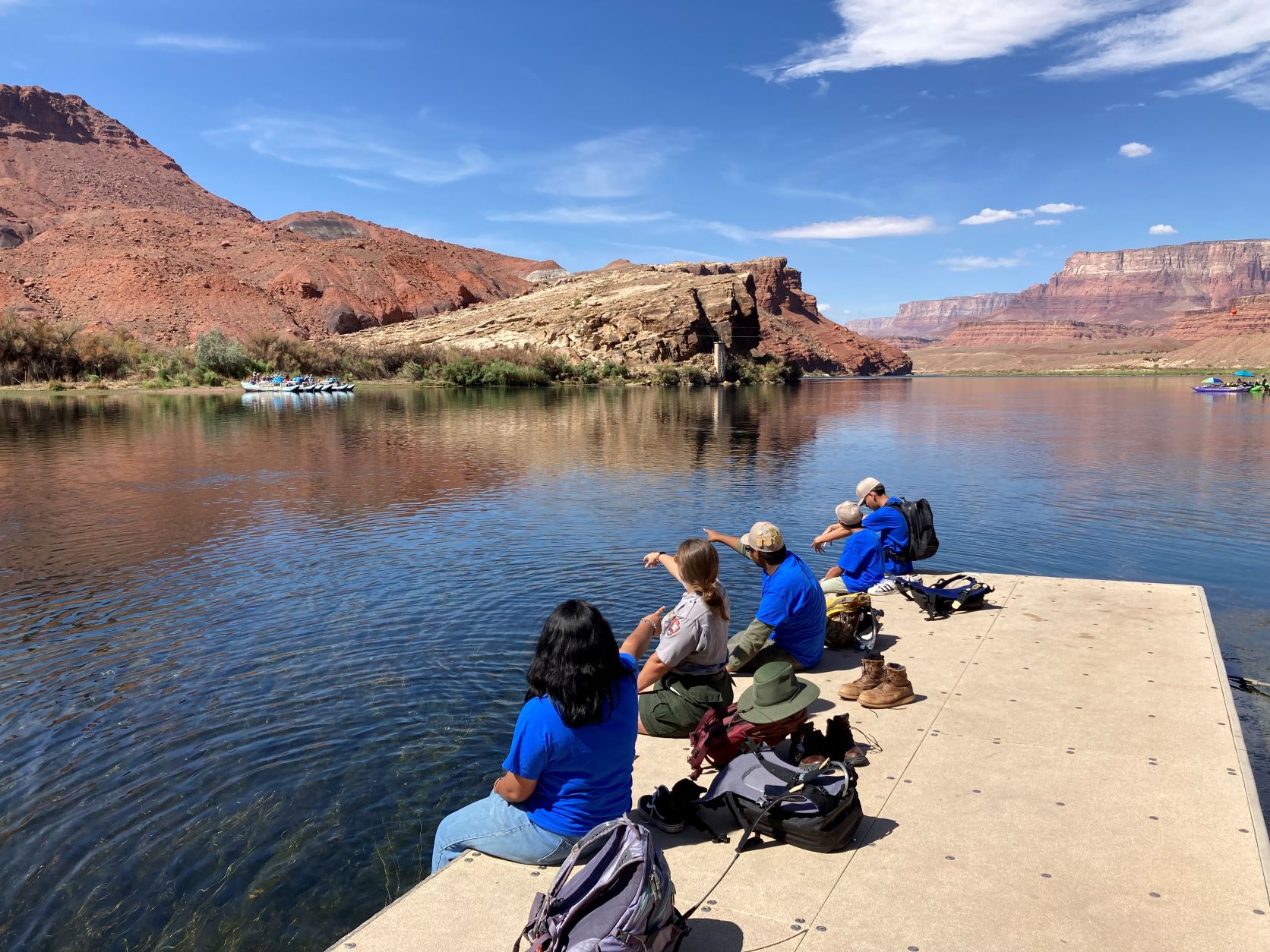 A park ranger sits with children on a river dock with tall canyon walls in the distance.