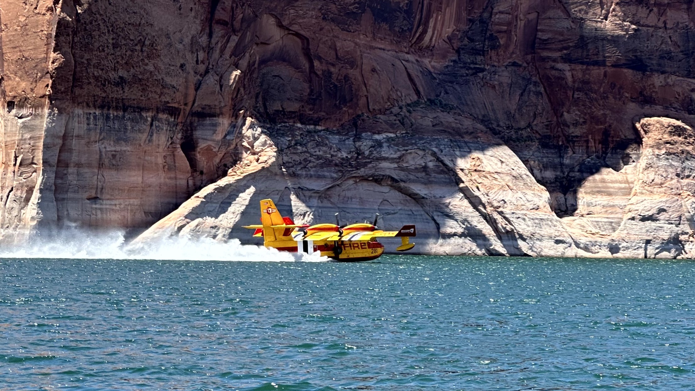A yellow and red plane scoops water from a lake in front of red rock cliffs.