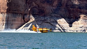 A yellow and red plane scoops water from a lake in front of red rock cliffs.