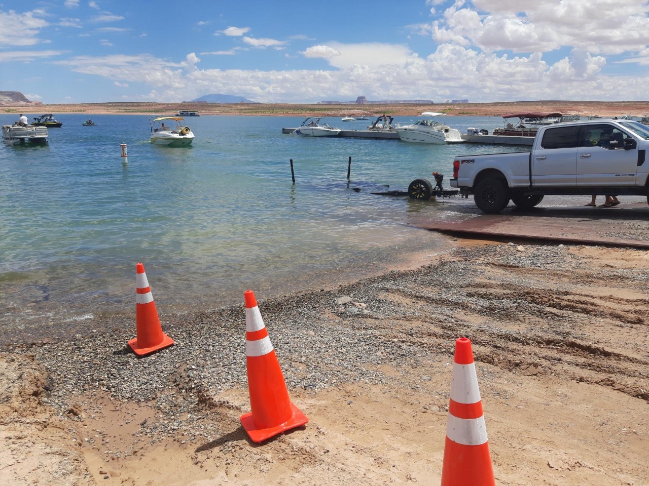 A truck pulling a trailer sits on the edge on metal ramp extension along waters edge.