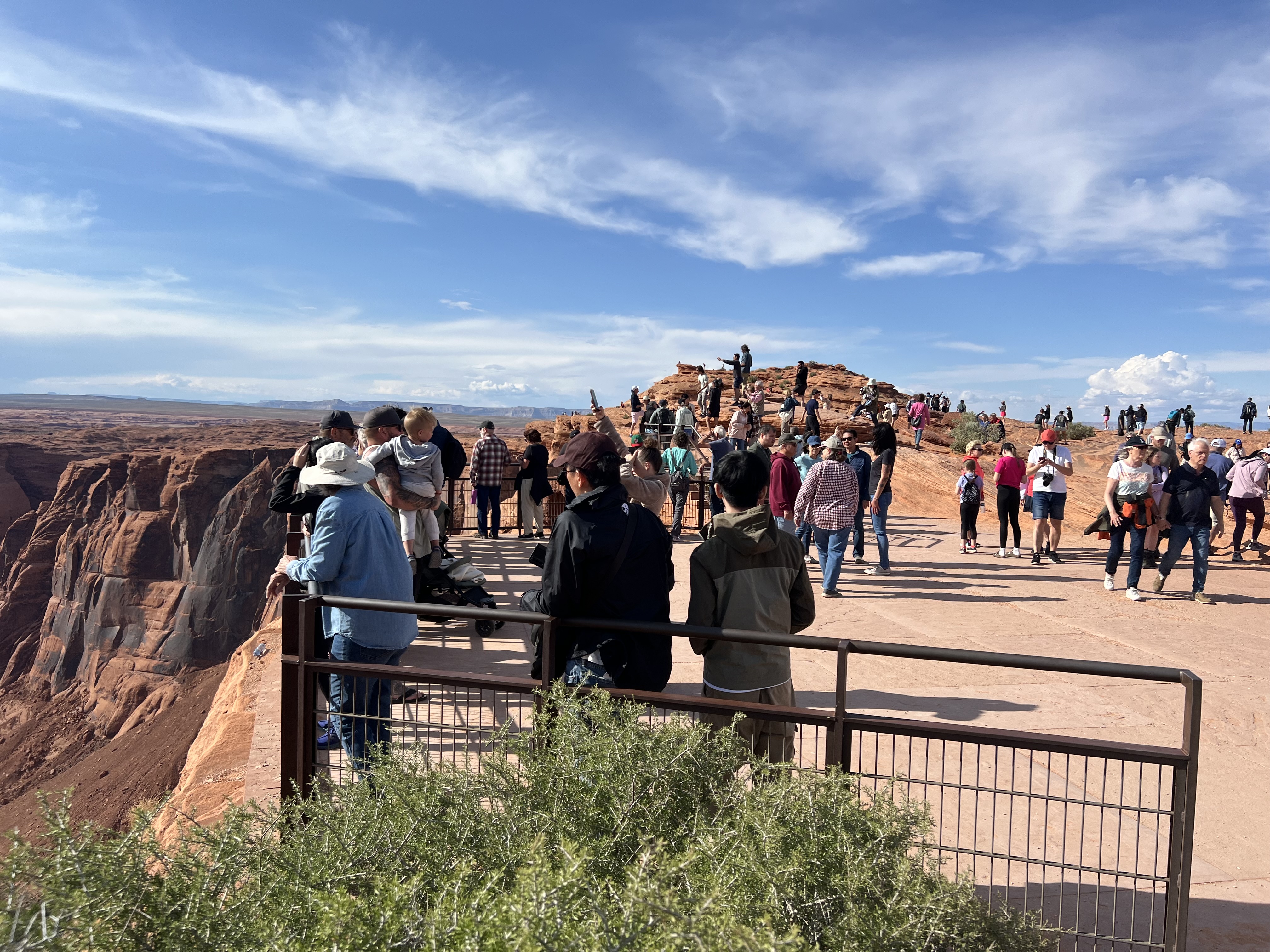 Railings at Horseshoe Bend Overlook a large group of people standing all over the rocks and surfaces behind a metal railing at a cliff.