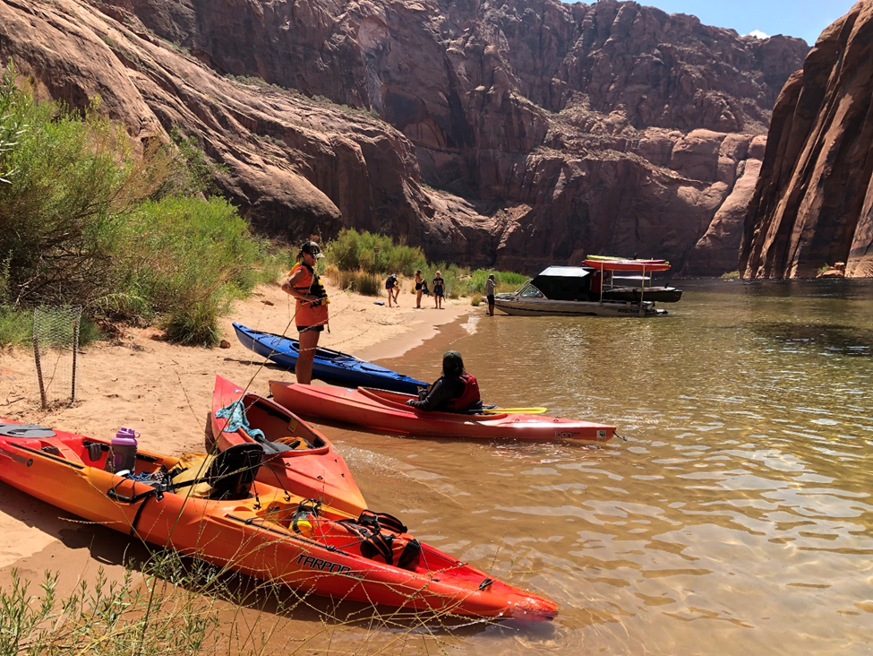 Half a dozed beached kayaks and people on a river in a sandstone canyon.