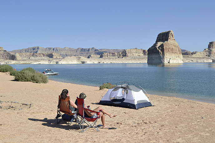 Two people sitting on a beach in front of a tent. in the water is a large rock structure. Cliffs in the background.