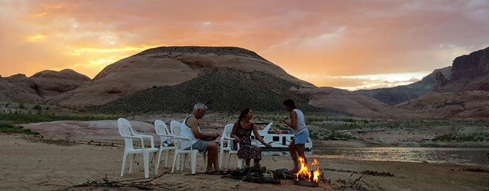 three people sit in plastic chairs around a campfire on a beach. Houseboat and sunset in background.