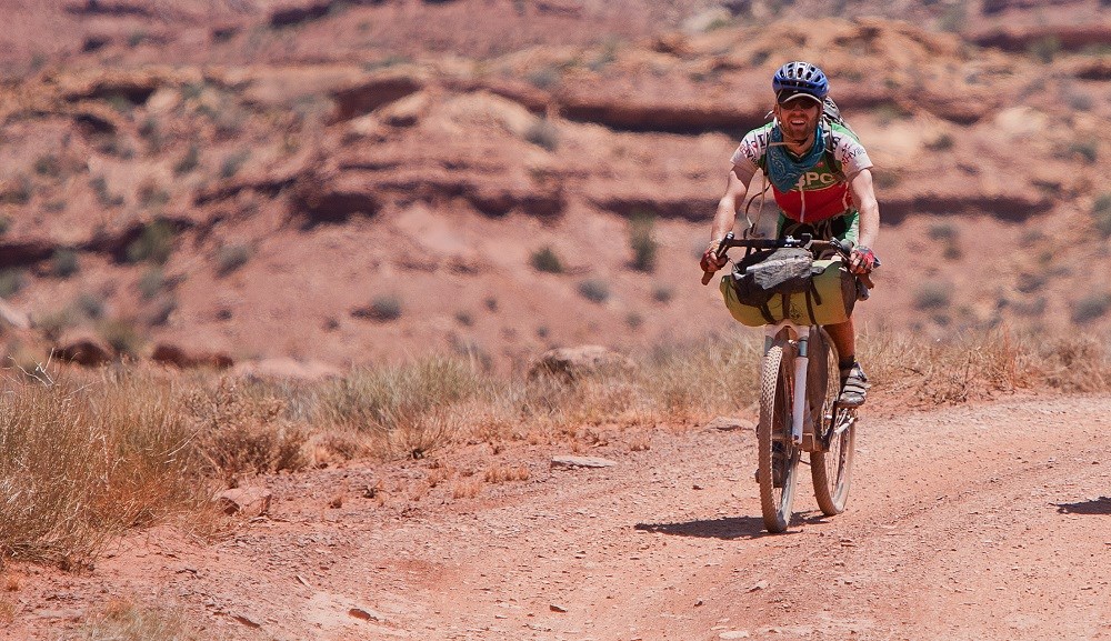 A smiling man with cycling gear rides a mountain bike on a dirt road in the desert
