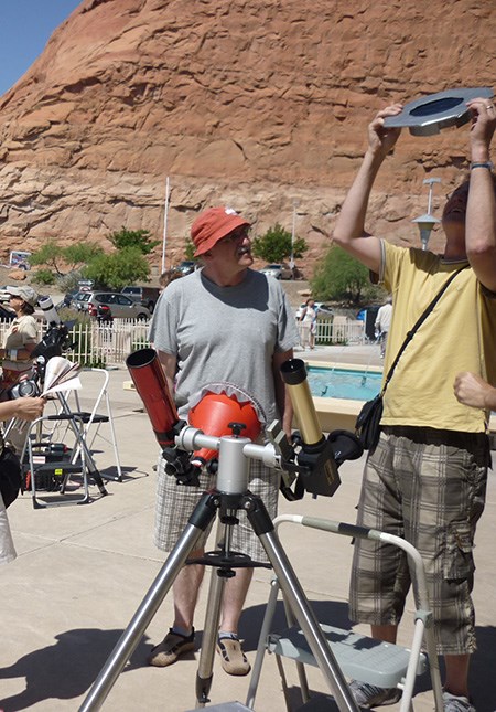 Two people stand next two solar telescopes. One holds up filter over his head and looks at the sun.