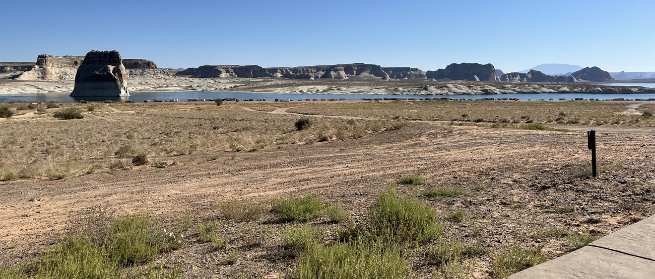 Dirt roads leads to a beach on a desert lake. Individuals are st up with shade shelters at the waters edge. A large sandstone butte sticks out to the water near the beach.