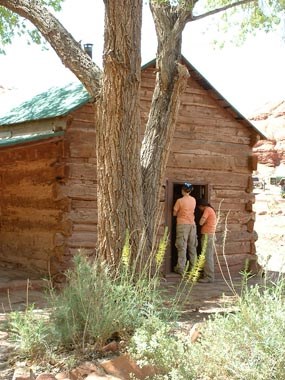 Two people stand in the doorway of a one-story wooden house.
