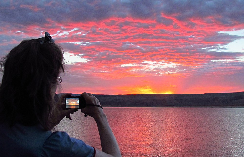 Woman photographs vibrantly colored clouds over lake. The image is repeated in her camera viewscreen. Woman photographs vibrantly colored clouds over lake. The image is repeated in her camera viewscreen.