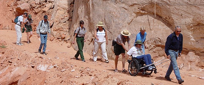 A group of people including two Park Rangers walks down a dirt trail next to a cliff. The person in the lead pulls a wheelchair while the person in the wheelchair operates the wheels and one of the Park Rangers pushes the chair from behind.