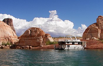 Houseboat on lakeshore with red rock formations and growing clouds in background