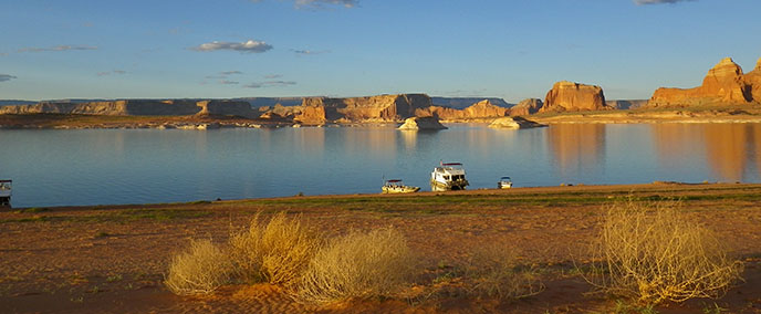 A panoramic view of Lake Powell with cliffs in the background and tumbleweeds in the foreground. In the middle are three boats beached. A panoramic view of Lake Powell with cliffs in the background and tumbleweeds in the foreground. In the middle are three boats beached.