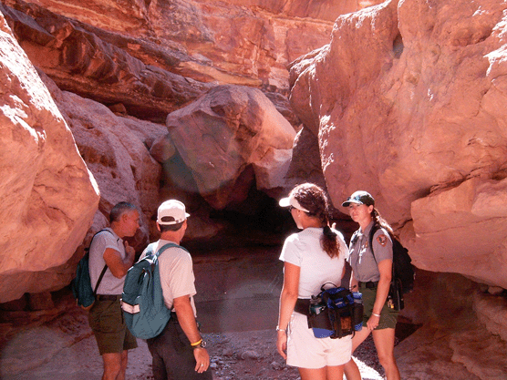 Four people stand at the entrance of a narrow canyon. They are all wearing backpacks or fanny packs. One of them is a park ranger. Four people stand at the entrance of a narrow canyon. They are all wearing backpacks or fanny packs. One of them is a park ranger.