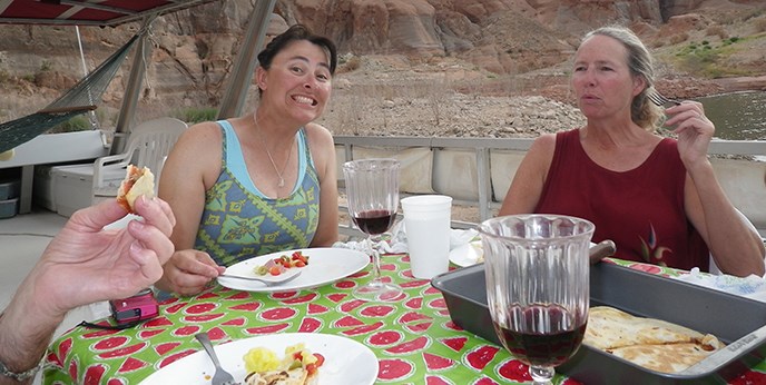 Two women sit at a table eating and smiling. A hand holding food is in the foreground. Two women sit at a table eating and smiling. A hand holding food is in the foreground.
