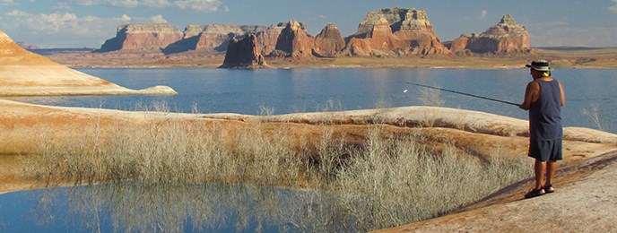 Man with fishing pole stands near reedy shore with red mesas and lake in background Man with fishing pole stands near reedy shore with red mesas and lake in background