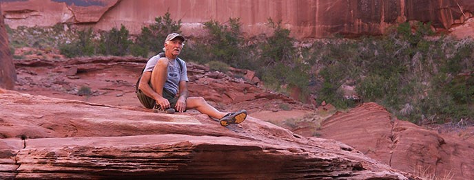 A man adjusts his shoe while sitting on the edge of a cliff. He is wearing a hat and a backpack. A man adjusts his shoe while sitting on the edge of a cliff. He is wearing a hat and a backpack.
