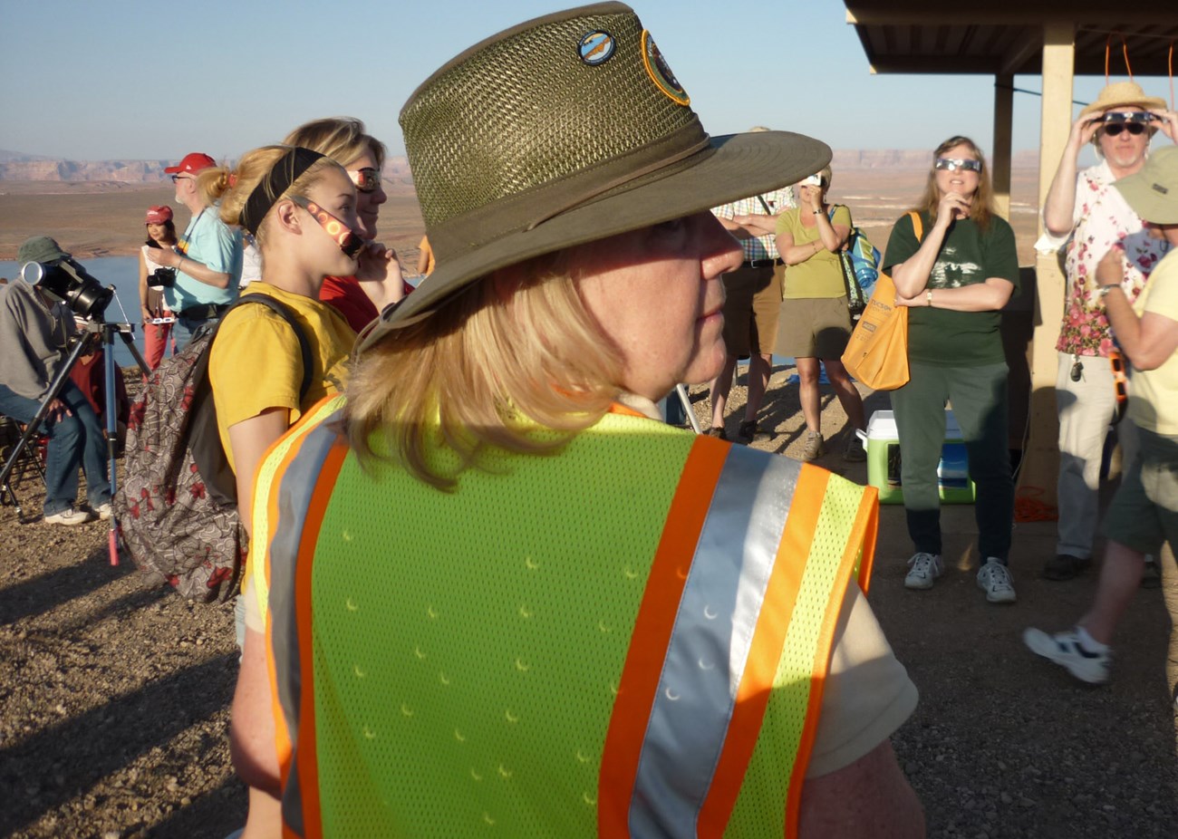 Park Ranger with images of a partial eclipse projected onto her reflective safety vest. People in background use eclipse glasses to view the sky.