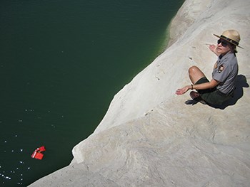 A ranger kneels at a cliff edge with tiny life jacket visible floating in water far below
