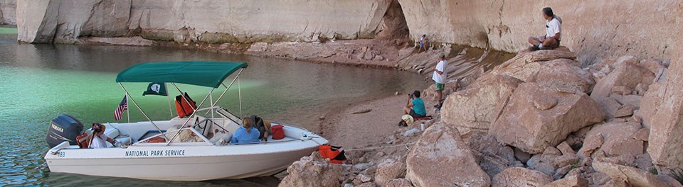 A white boat labeled "National Park Service" is beached in a sandstone alcove. Lifejackets and gear are on board. Four people are on the beach. A white boat labeled "National Park Service" is beached in a sandstone alcove. Lifejackets and gear are on board. Four people are on the beach.