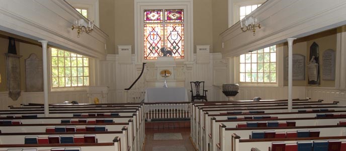 Color photo showing church interior with two sections of pews separated by a center aisle and facing the altar which is situated just below a large multi-colored stained glass window. Color photo showing church interior with two sections of pews separated by a center aisle and facing the altar which is situated just below a large multi-colored stained glass window.