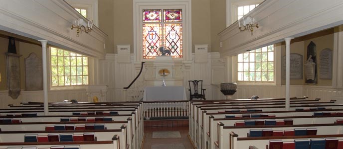 Color photo showing church interior with two sections of pews separated by a center aisle and facing the altar which is situated just below a large multi-colored stained glass window.